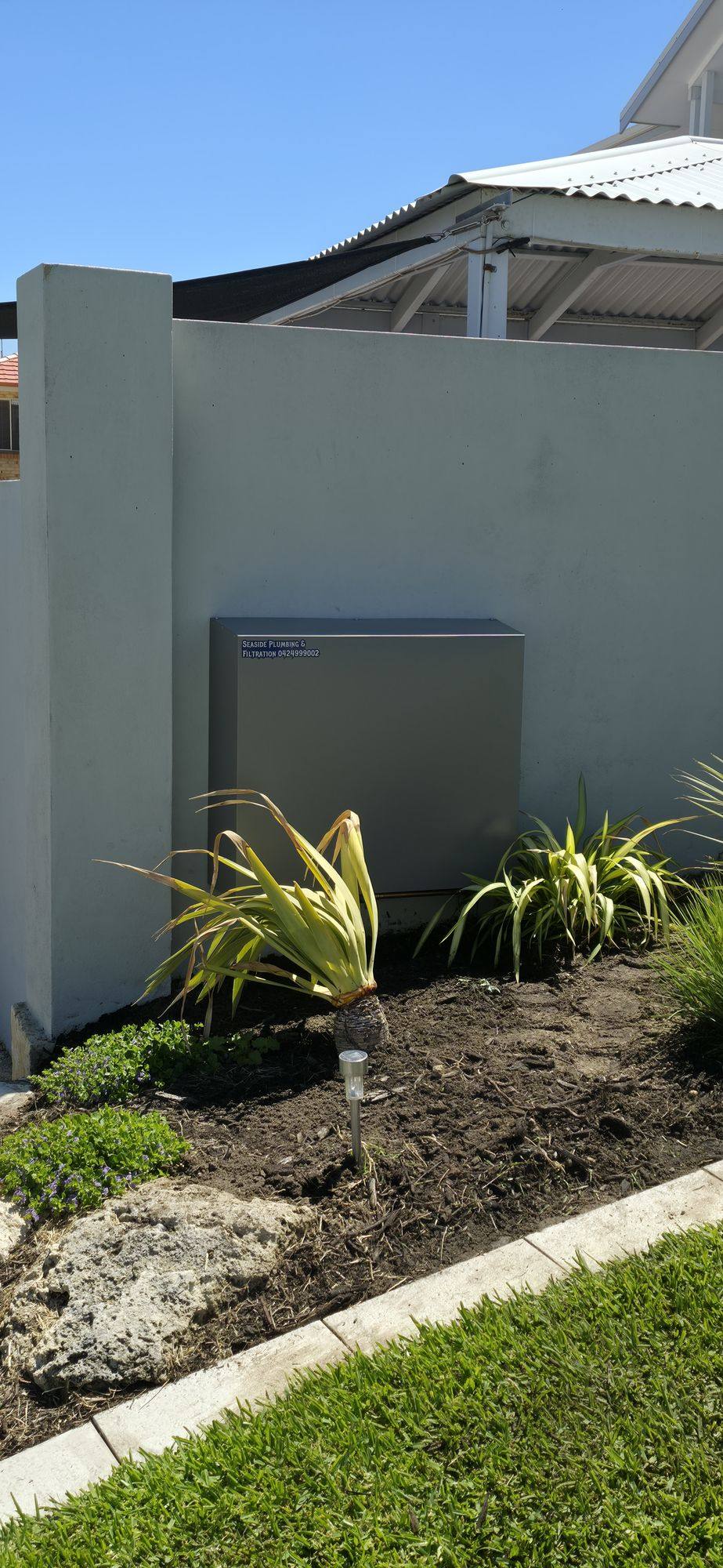 A sage-grey filter cover on a rendered garden wall above a planted bed