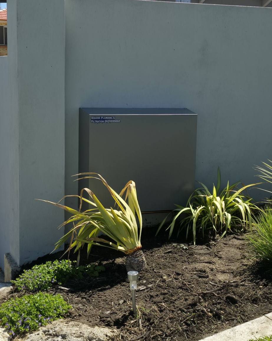 A sage-grey filter cover on a rendered garden wall above a planted bed