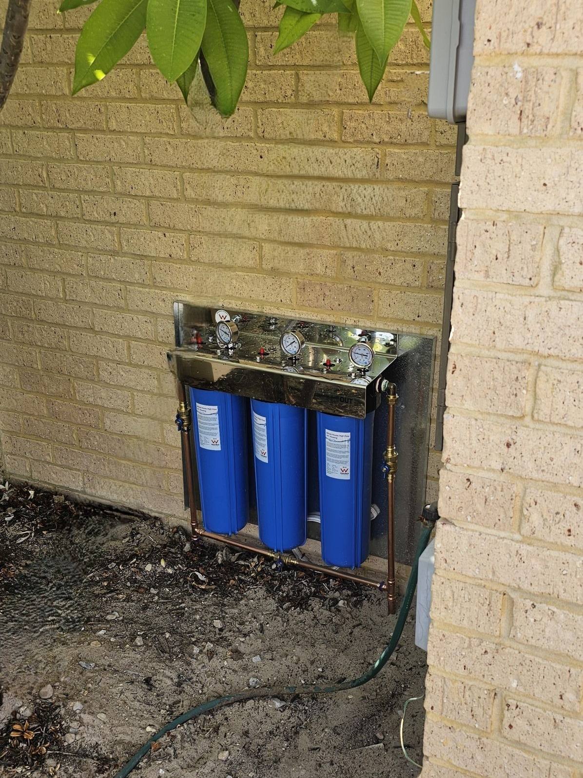 A three-stage whole-house filter with blue housings on a limestone-brick wall beside a frangipani
