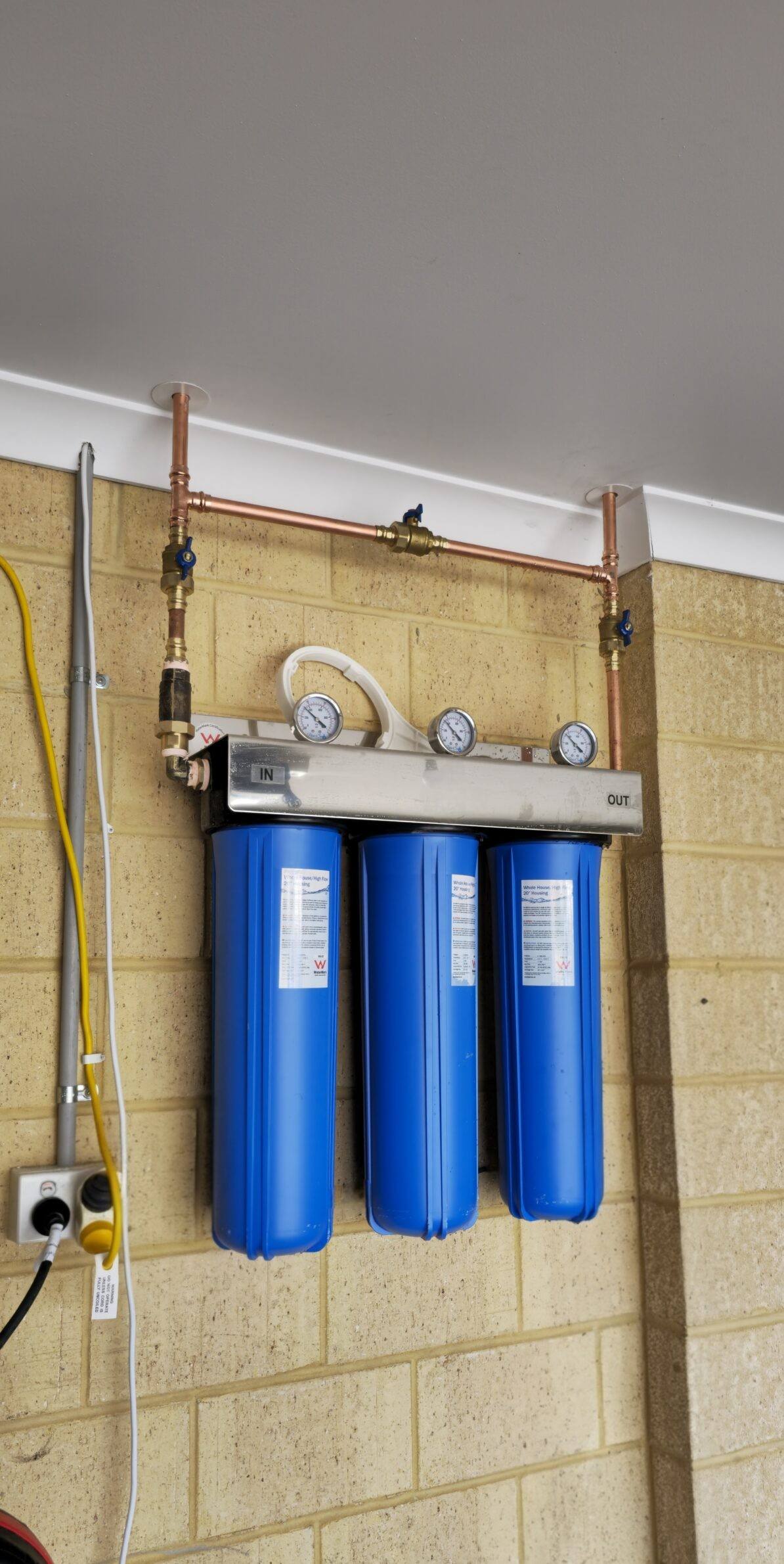 Three large blue filter housings on a stainless manifold with copper pipe, mounted on a garage wall