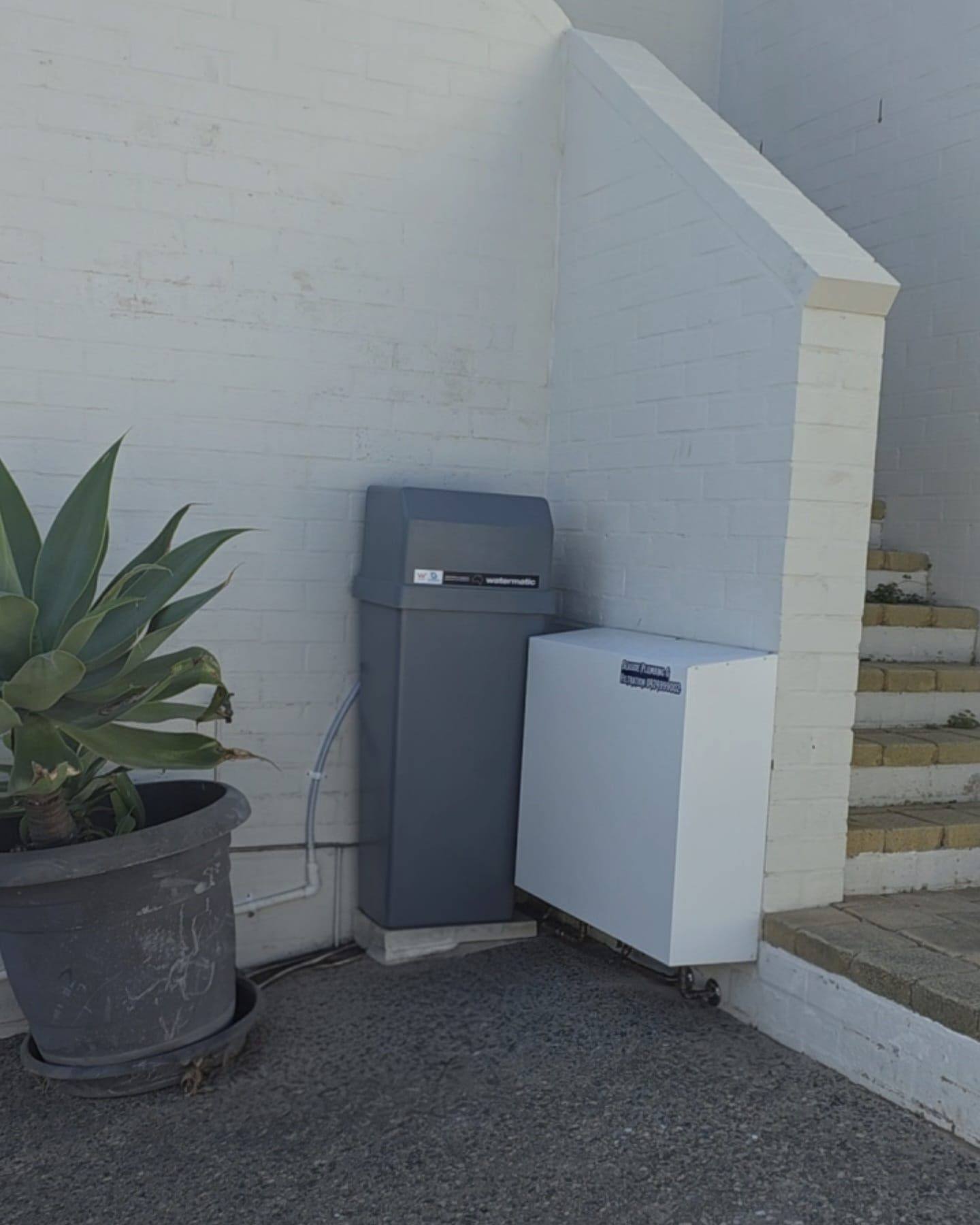 An outdoor water softener and a white filter cover beside a painted brick wall