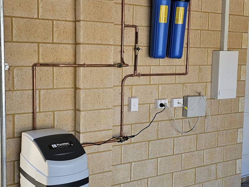 A floor-standing Puretec water softener connected to two blue filter housings on a garage wall