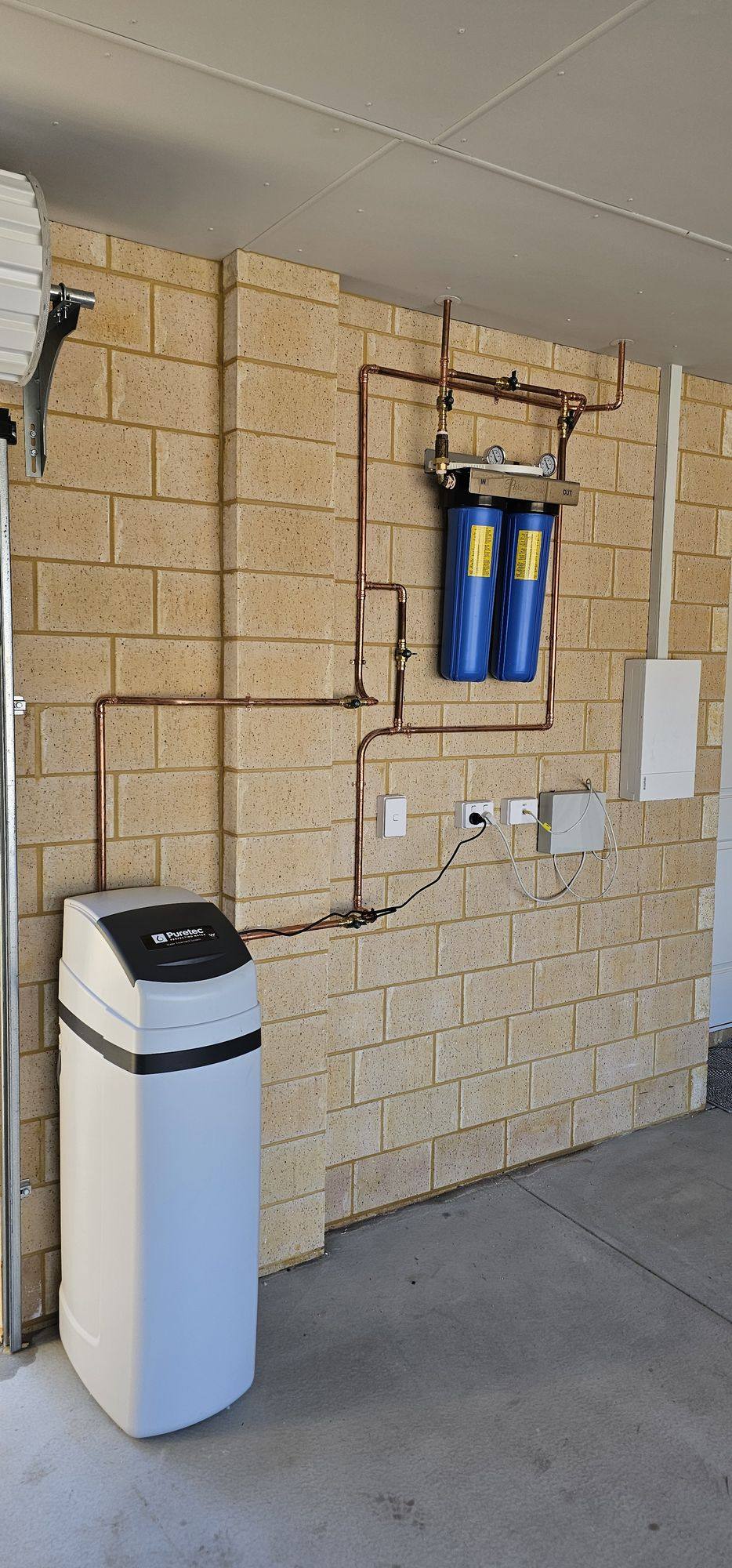 A floor-standing Puretec water softener connected to two blue filter housings on a garage wall