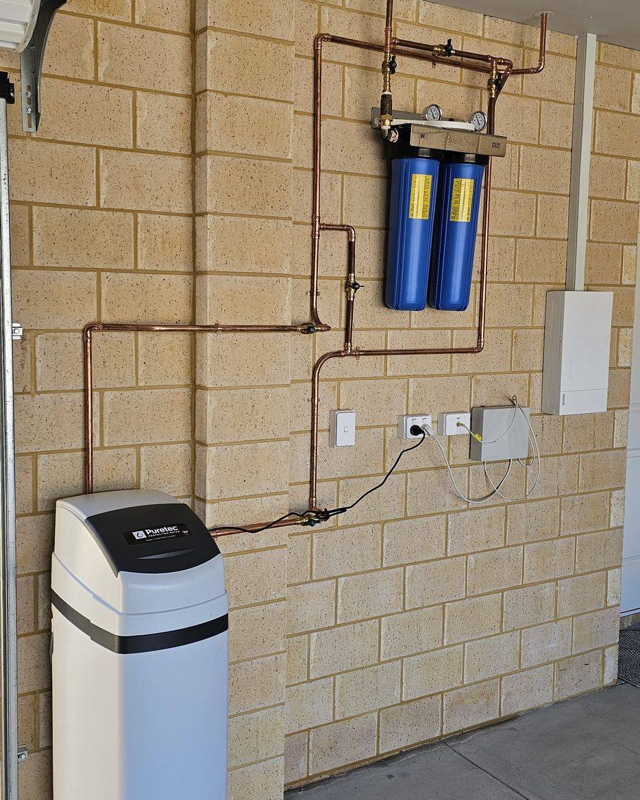 A floor-standing Puretec water softener connected to two blue filter housings on a garage wall