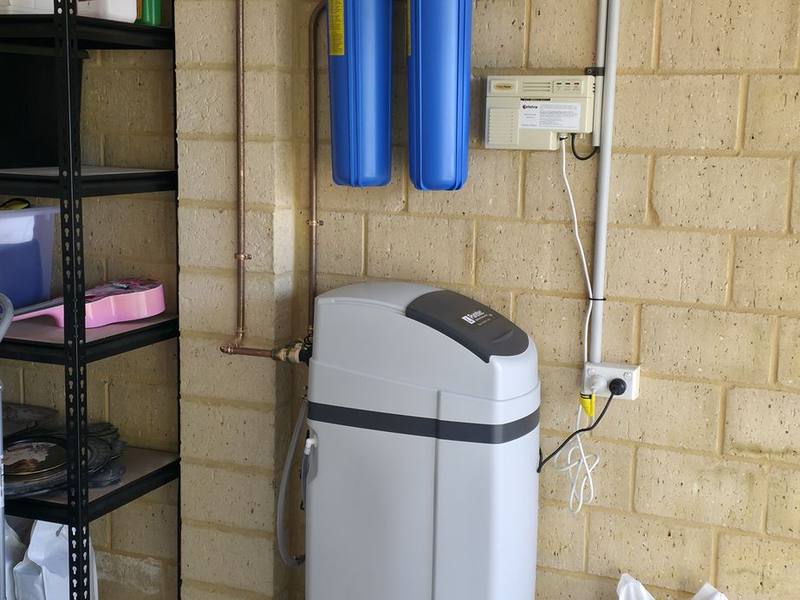A water softener below two blue filter housings, with a bag of softener salt alongside