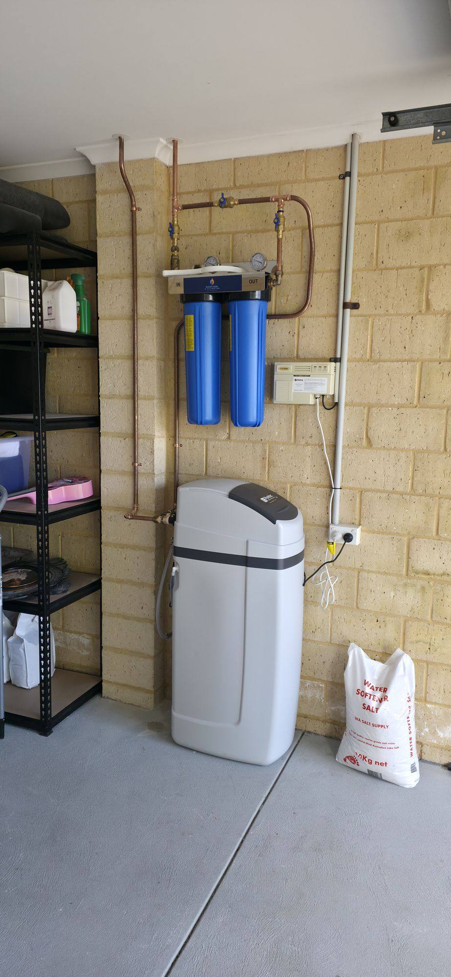 A water softener below two blue filter housings, with a bag of softener salt alongside