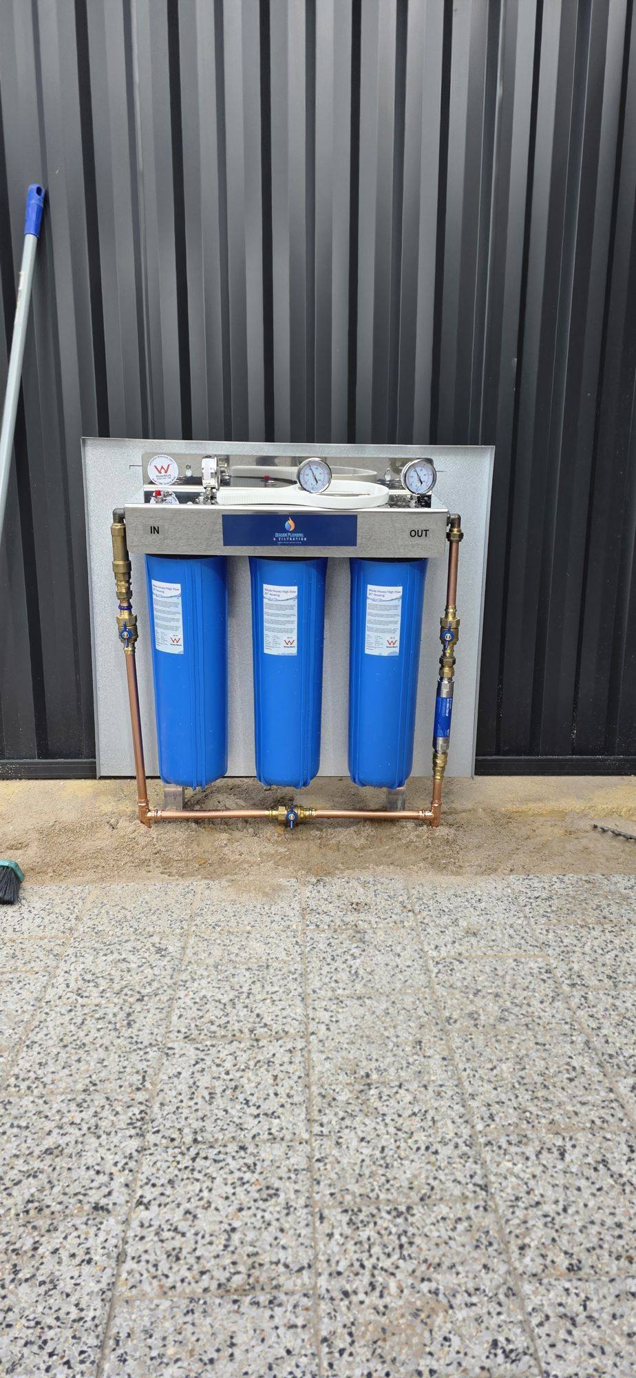 Three blue filter housings with copper pipework on a backing panel beside a dark fence