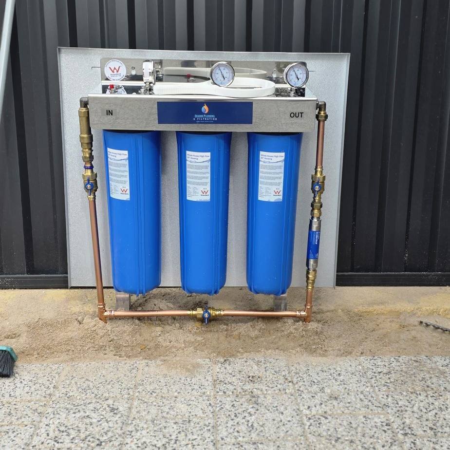 Three blue filter housings with copper pipework on a backing panel beside a dark fence