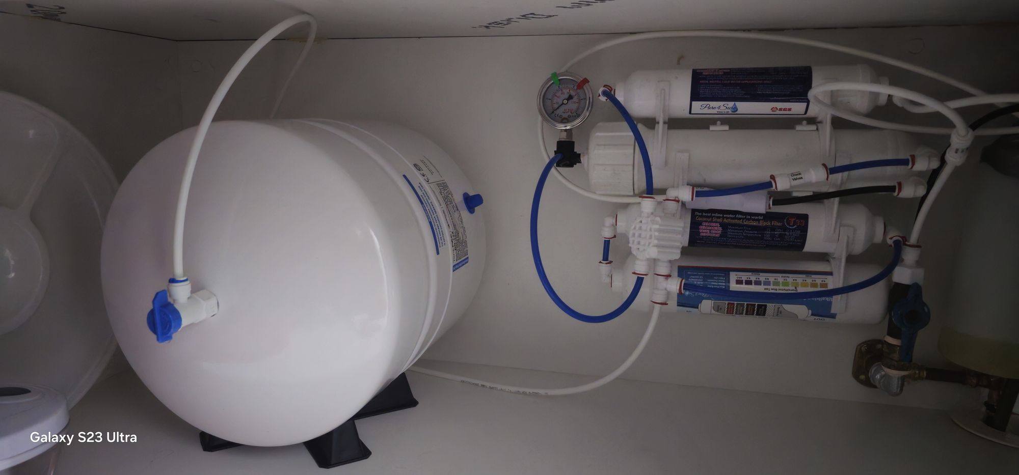Reverse osmosis filter stages and tubing beside a horizontal white storage tank inside a cupboard