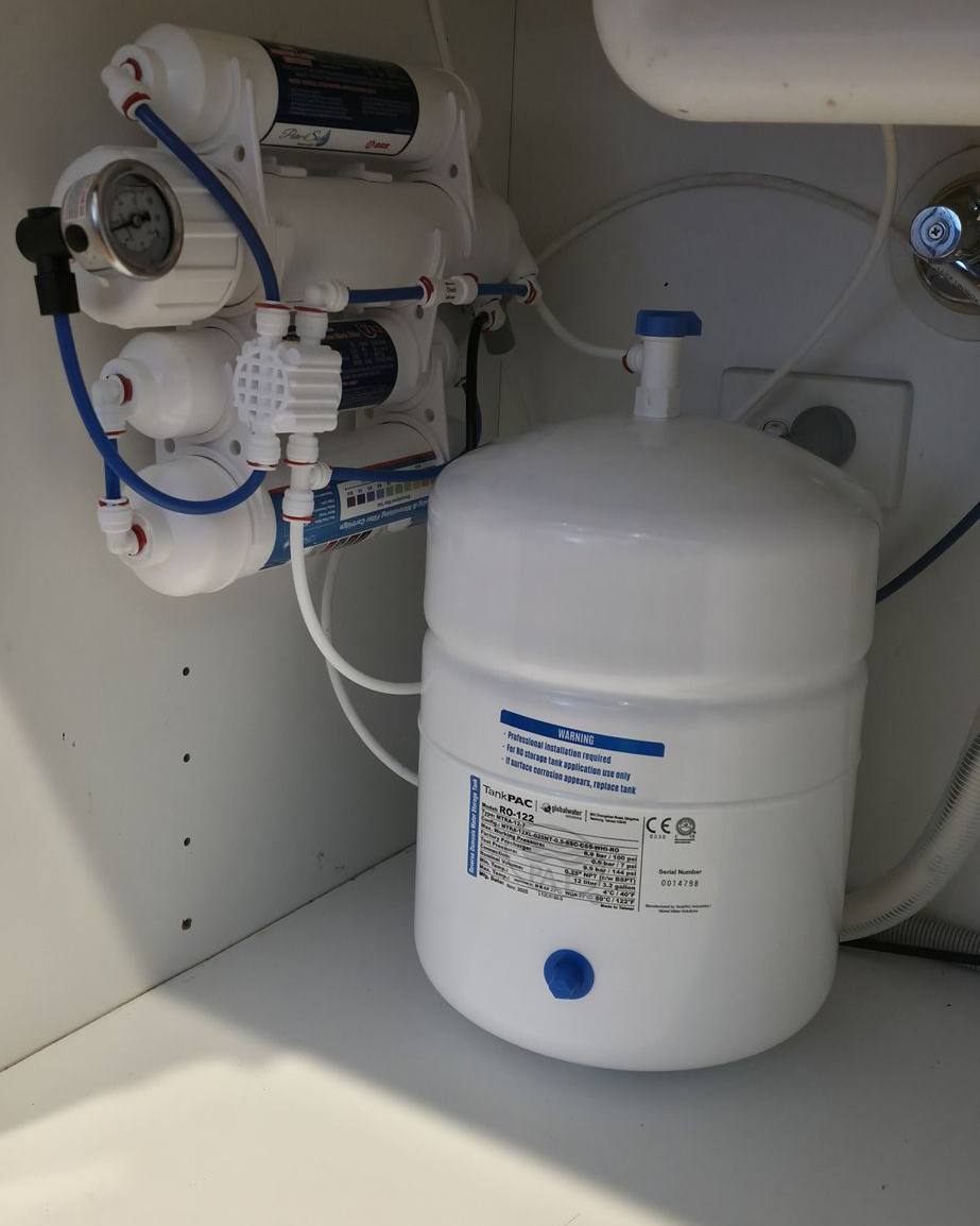 A reverse osmosis filter assembly and white storage tank in the cupboard beneath a kitchen sink